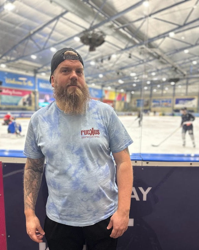 Blue tie-dye t shirt with burgundy ruckus logo to left chest, being worn by one of our ambassadors, Martyn. Background of the image is the ice rink and boards.