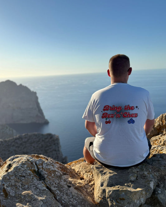 Person wearing a t shirt with Bring the Bez n Chez on, sitting on a rocky cliff overlooking the ocean with a scenic view.